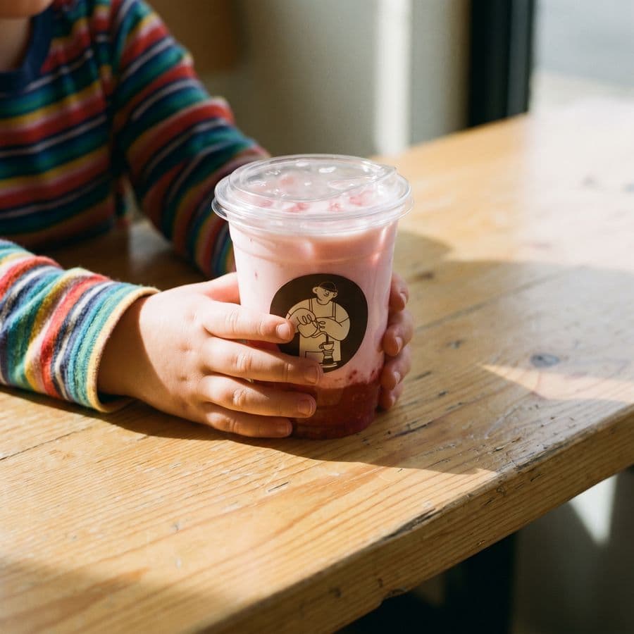 A child's hands holding a cup of strawberry milk on a wooden table