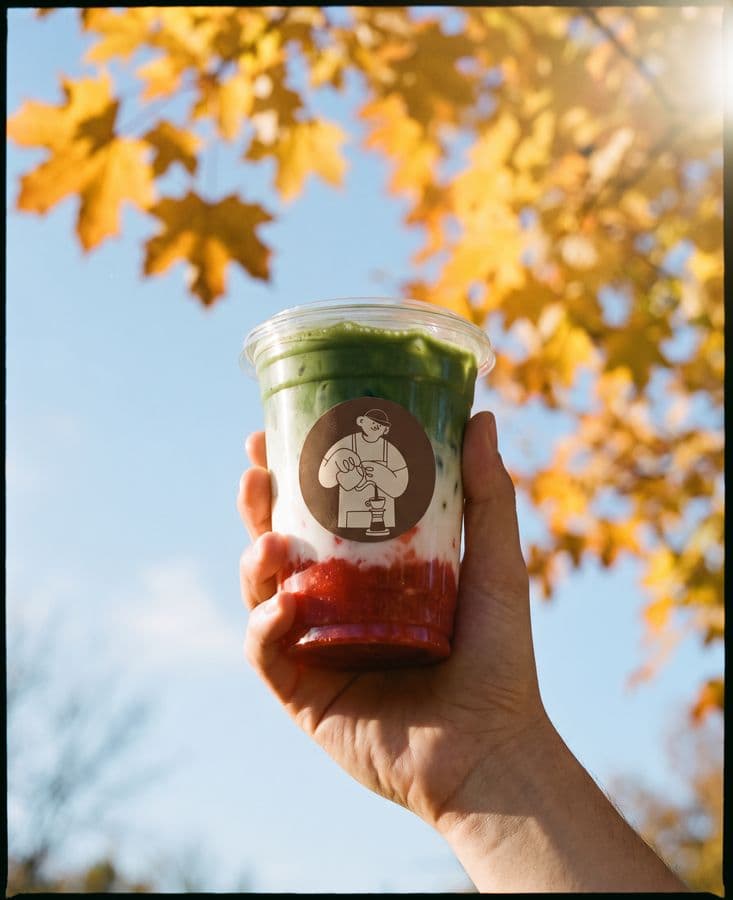 An iced strawberry matcha latte held up against an autumn sky